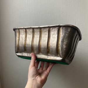 May include: A rectangular metal planter with vertical gold-colored stripes and a hammered texture. The planter has a dark, aged appearance and a green felt base. The planter is held by a hand.