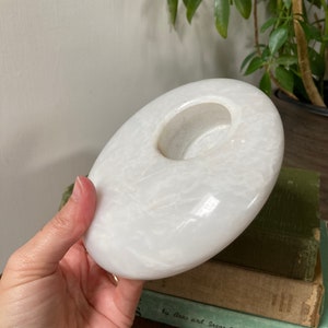 May include: White marble candle holder with a round, slightly concave top and a circular opening in the centre. The holder has a smooth, polished surface with subtle veining.