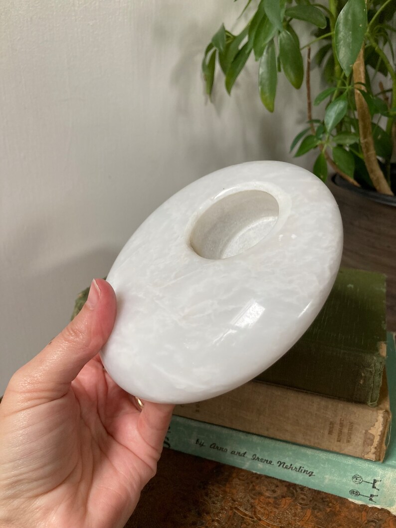 May include: White marble candle holder with a round, slightly concave top and a circular opening in the centre. The holder has a smooth, polished surface with subtle veining.