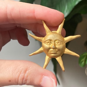 May include: A gold-colored sun-shaped brooch with a smiling face and pointed rays. The brooch is held in a person's hand against a blurred background of green foliage and a blue surface.