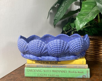 May include: A blue ceramic planter shaped like seashells, sitting on top of a stack of three books. The books are titled "The most important thing in the world", "Better Flower Arrangements", and "Gardening with Perennials Month by Month".