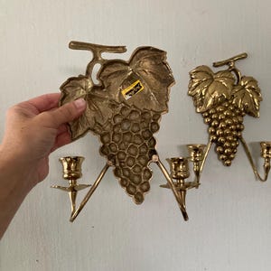 May include: Two brass wall sconces shaped like grape clusters with leaves. Each sconce has three candle holders. One sconce is being held by a hand. The sconces are mounted on a light-colored wall.
