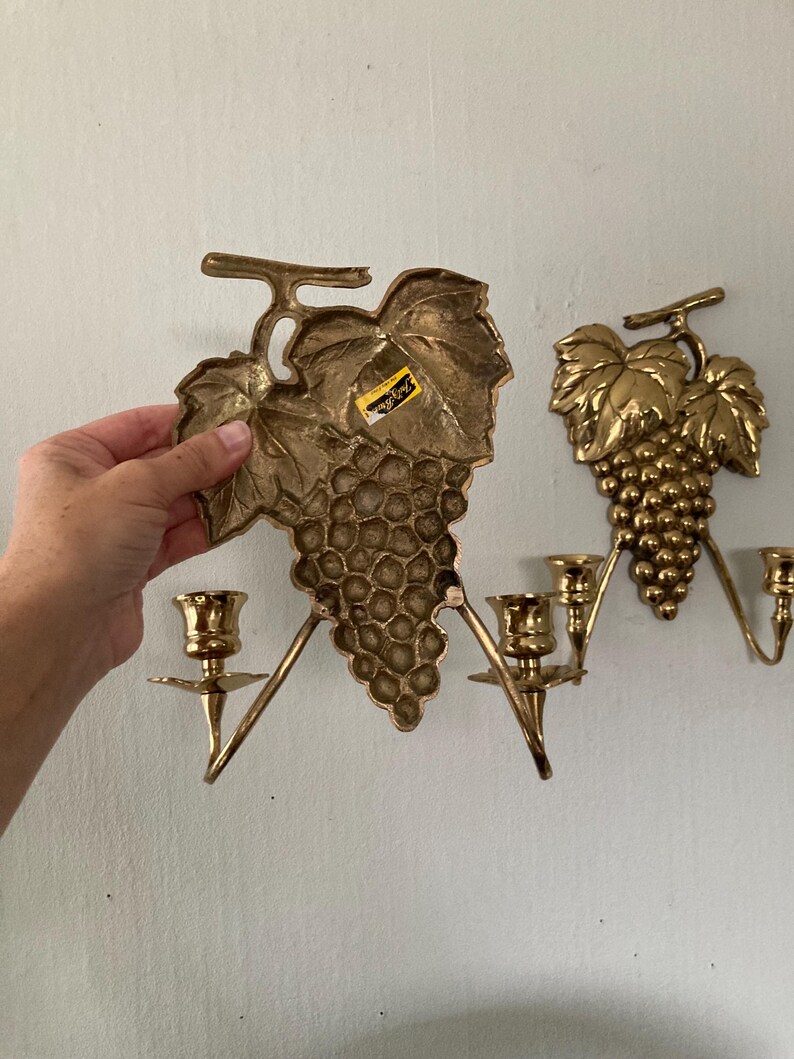 May include: Two brass wall sconces shaped like grape clusters with leaves. Each sconce has three candle holders. One sconce is being held by a hand. The sconces are mounted on a light-colored wall.