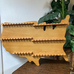 May include: Wooden wall shelf shaped like the United States of America with multiple slots for displaying items. The shelf is made of light brown wood and is mounted on a wall.