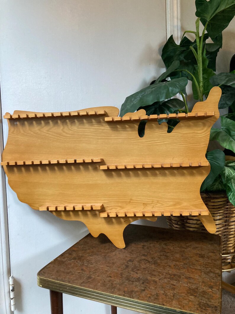 May include: Wooden wall shelf shaped like the United States of America with multiple slots for displaying items. The shelf is made of light brown wood and is mounted on a wall.