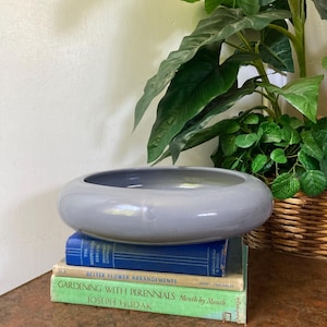 May include: A smooth, light gray ceramic bowl with a wide, rounded rim sits atop a stack of three books. The books include titles such as The Deepening Stream, Better Flower Arrangements, and Gardening with Perennials by Joseph Hudak.