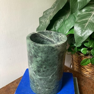 May include: A cylindrical green stone vessel with a polished finish, featuring natural dark green and black mottled patterns and white veining throughout the surface. The object has a smooth, rounded rim and a hollow interior, standing upright.