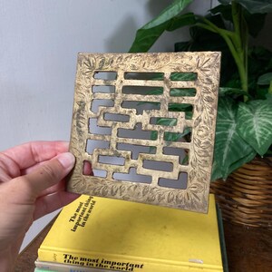 May include: A square, gold-colored metal decorative grate with a floral design around the edges. The grate is held above a stack of books with the titles "The most important thing in the world" and "Gardening with Perennials".