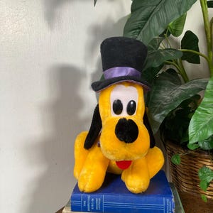 May include: A plush yellow Pluto dog toy wearing a black top hat with a purple band. The toy has black ears, a black nose, and a red tongue. It sits atop a stack of vintage books with titles like "Better Flower Arrangements."