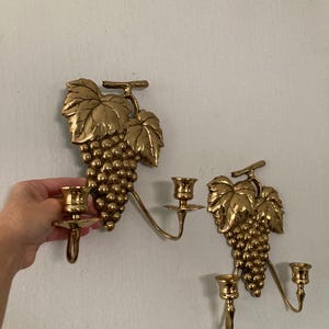 May include: Two brass wall sconces, each featuring a cluster of grapes and leaves. The sconces have two candle holders each, with a curved arm extending from the grape cluster. The sconces are mounted on a white wall.