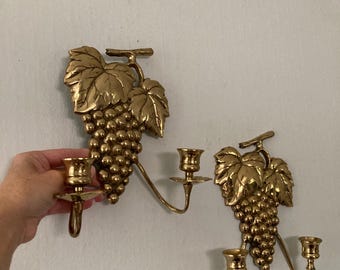 May include: Two brass wall sconces, each featuring a cluster of grapes and leaves. The sconces have two candle holders each, with a curved arm extending from the grape cluster. The sconces are mounted on a white wall.