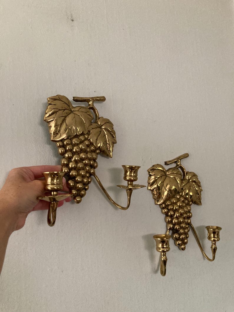 May include: Two brass wall sconces, each featuring a cluster of grapes and leaves. The sconces have two candle holders each, with a curved arm extending from the grape cluster. The sconces are mounted on a white wall.