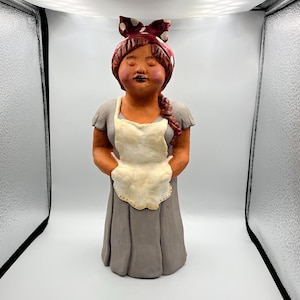 Può includere: Una statuetta in ceramica di una donna che indossa un vestito grigio e un grembiule bianco. Ha una fascia per capelli a pois rossi e bianchi e gli occhi chiusi.