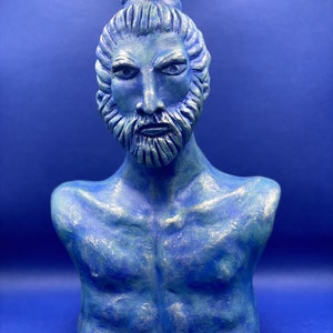 Könnte beinhalten: Blaue Büstenskulptur eines Mannes mit Bart und zu einem Dutt gebundenem Haar. Die Skulptur hat eine strukturierte Oberfläche mit silbernen Akzenten. Der Hintergrund ist einfarbig blau.