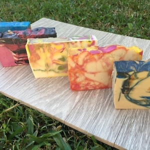 May include: A selection of colourful handmade soaps on a light grey wooden surface. The soaps display a variety of vibrant colours and swirling patterns, including blue, pink, yellow, and orange. The soaps are rectangular and appear freshly cut.