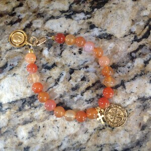 May include: A gold bracelet with a St. Benedict medal charm and carnelian beads in shades of orange and pink.