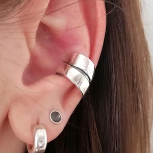 Könnte beinhalten: Ein silberner Ohrring mit einem Doppelband-Design. Der Ohrring wird am oberen Teil des Ohrs getragen. Ein kleiner silberner Creolen-Ohrring wird im unteren Ohrläppchen getragen. Ein kleiner schwarzer Ohrstecker wird im Ohrknorpel getragen.