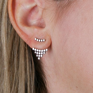 Puede incluir: Un par de pendientes de plata con pequeñas piedras transparentes. Los pendientes tienen forma de triángulo y barra curva.