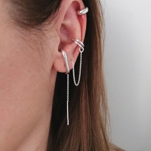 Könnte beinhalten: Ein silberner Ohrring mit einer Kette, die herunterhängt. Der Ohrring wird am oberen Teil des Ohrs getragen, und die Kette hängt bis zum unteren Teil des Ohrläppchens herunter. Der Ohrring ist aus glattem, glänzendem Metall.