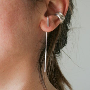 Op de afbeelding: Een zilveren oorcuff met een zilveren oorbel. De oorbel is een lange, dunne staaf die van het oor hangt.