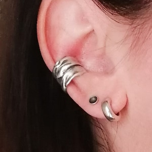 May include: A silver ear cuff with a twisted design and a silver hoop earring. The earrings are worn in the left ear.