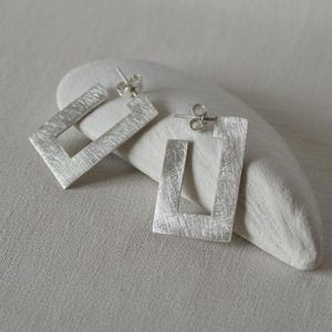 May include: A pair of silver-toned earrings featuring an open rectangular frame design with a brushed, textured finish. Each earring includes a standard post back closure, showcasing a clean, geometric shape with a matte metallic appearance.