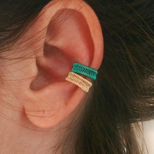 May include: A close-up of a person's ear wearing two ear cuffs. The top cuff is a bright teal color and the bottom cuff is a gold color.