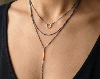 Puede incluir: Un conjunto de collares en capas con tres hilos. El hilo superior tiene un pequeño colgante de círculo dorado, el hilo del medio es una cadena simple y el hilo inferior tiene un colgante de barra dorada con una larga caída de cadena. Las cadenas son de color gris oscuro.