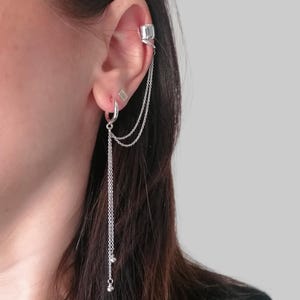 May include: Silver ear cuff and earring set. The ear cuff has a square design and connects to the earring via delicate chains. The earring is a hoop with a dangling chain and small silver beads. The set is worn on the ear.