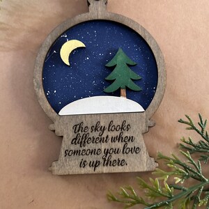 May include: Wooden Christmas ornament shaped like a snow globe. The interior features a dark blue night sky with a yellow crescent moon, a green tree, and white snow. The text "The sky looks different when someone you love is up there" is engraved on the bottom.