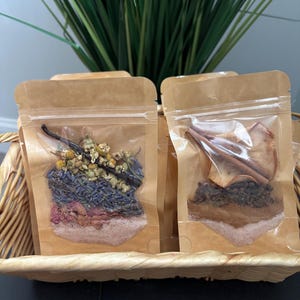 May include: Two clear pouches filled with dried herbs and spices, displayed in a woven basket. One pouch contains lavender, chamomile, and a vanilla bean. The other contains cinnamon sticks and dried apple slices. Green foliage is visible in the background.