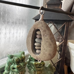 May include: A unique, handcrafted stone pendant with a hollowed-out center, holding several small, smooth stones. The pendant is suspended by a silver wire loop. The background includes a decorative lantern and ceramic figurines.
