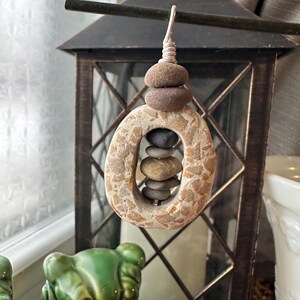 May include: A handcrafted stone wind chime. The chime features a stack of small, smooth stones suspended from a wire. The main body is an oval-shaped stone with a hole in the center, and a stack of stones inside.