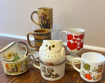 May include: A collection of vintage ceramic mugs with various designs. The mugs feature a brown and white owl, a red heart pattern, a brown and white striped design, a brown and white floral design, and a white mug with a green and brown floral design.