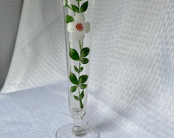 May include: A clear glass vase with a white flower and green leaves painted on the side. The vase has a flared top and a round base.