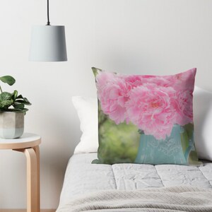 May include: A bouquet of pink peonies in a light blue ceramic pitcher. The pitcher has a floral design and is sitting on a white bedspread.
