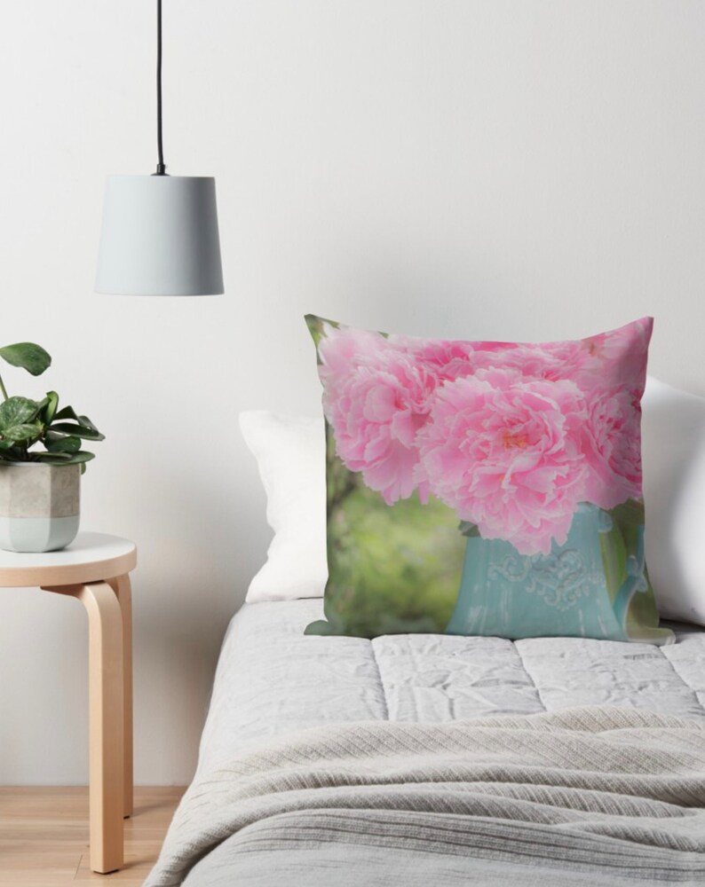 May include: A bouquet of pink peonies in a light blue ceramic pitcher. The pitcher has a floral design and is sitting on a white bedspread.