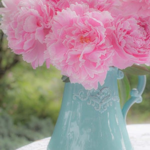 May include: A bouquet of pink peonies in a light blue ceramic pitcher with a decorative design. The pitcher is sitting on a white lace tablecloth.