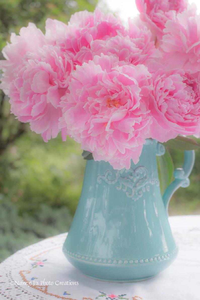 May include: A bouquet of pink peonies in a light blue ceramic pitcher with a decorative design. The pitcher is sitting on a white lace tablecloth.