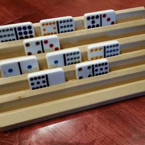 May include: A wooden domino rack with a set of dominoes. The rack has seven slots for dominoes and is made of light wood. The dominoes are white with black dots and some have colored dots. The dominoes are arranged in a pattern on the rack.