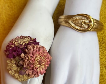 May include: A gold-tone bracelet with a large, ornate buckle. The bracelet is on a white mannequin hand.