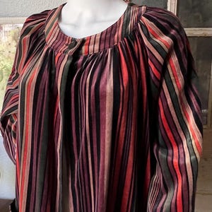 May include: A multi-colored striped top with long sleeves. The vertical stripes include shades of red, black, purple, and green. The top has a round neckline and a single button closure. The fabric appears to be a soft, flowing material.