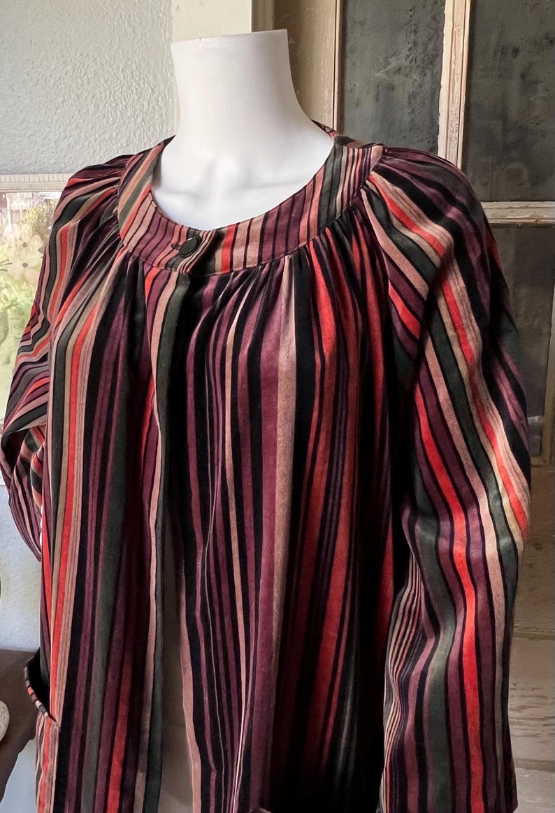 May include: A multi-colored striped top with long sleeves. The vertical stripes include shades of red, black, purple, and green. The top has a round neckline and a single button closure. The fabric appears to be a soft, flowing material.
