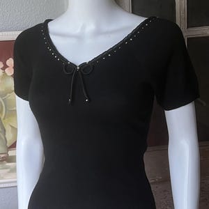 May include: A black short-sleeved knit top featuring a V-neckline adorned with small silver-toned studs and a decorative black bow at the center. The garment has a fitted silhouette and is displayed on a white mannequin torso.