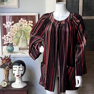 May include: A long-sleeved, open-front jacket with vertical stripes in shades of red, black, and brown. The jacket has two front pockets and is displayed on a mannequin. The jacket's design is vintage-inspired.