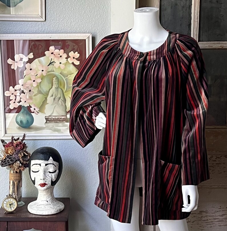 May include: A long-sleeved, open-front jacket with vertical stripes in shades of red, black, and brown. The jacket has two front pockets and is displayed on a mannequin. The jacket's design is vintage-inspired.