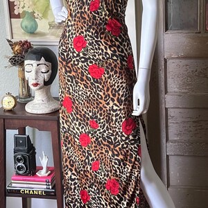 May include: A sleeveless, floor-length dress featuring a leopard print pattern with red rose accents. The dress has a keyhole neckline and a high slit on one side. The dress is displayed on a mannequin.