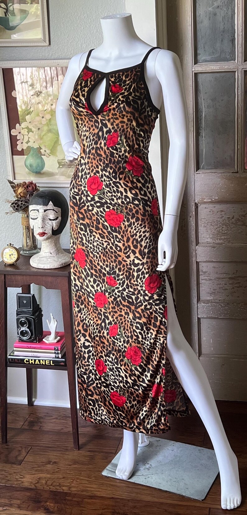 May include: A sleeveless, floor-length dress featuring a leopard print pattern with red rose accents. The dress has a keyhole neckline and a high slit on one side. The dress is displayed on a mannequin.