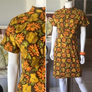 May include: A short-sleeved dress featuring a high neckline and a vibrant floral pattern in shades of orange, mustard yellow, olive green, and dark brown. The garment has a fitted bodice with a horizontal band at the waist and a straight skirt.