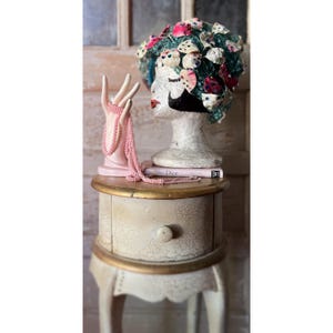 Puede incluir: Una exhibición decorativa con un soporte de joyas rosa en forma de mano y un collar de cuentas rosa. Una mesita de noche de estilo vintage sostiene una cabeza de maniquí adornada con un tocado floral y un libro con la palabra "Dior".